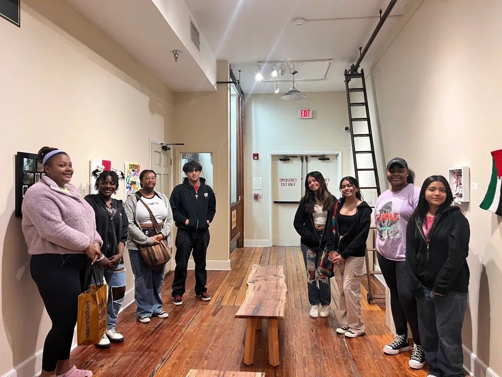 People standing in a gallery hall beside framed artwork, with benches and a ladder in the background