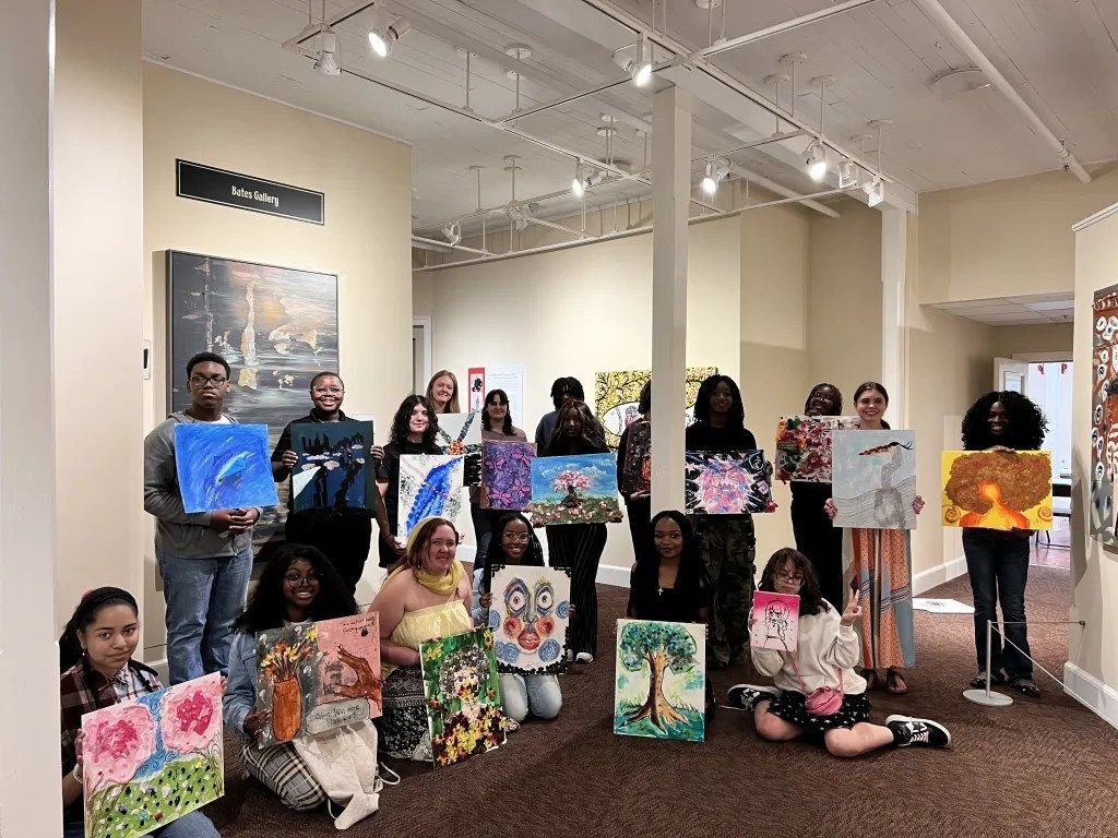 Group holding colorful paintings in an art gallery, standing and kneeling beneath bright lights.