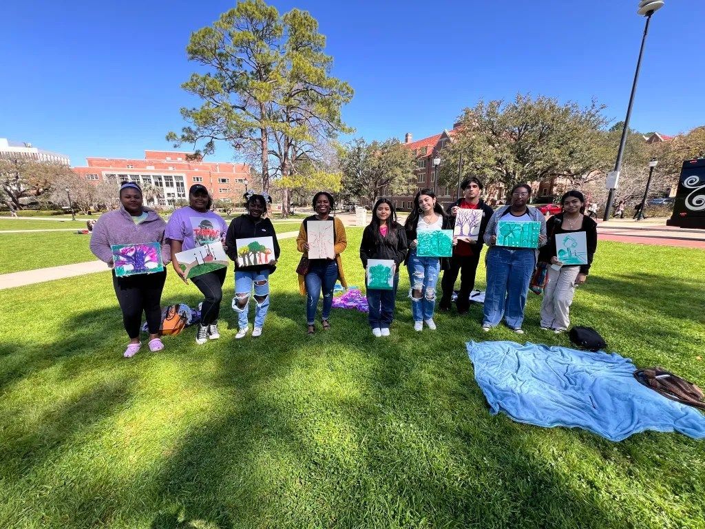 Group of people standing on a sunny lawn holding signs, with a blue blanket spread on the grass nearby.