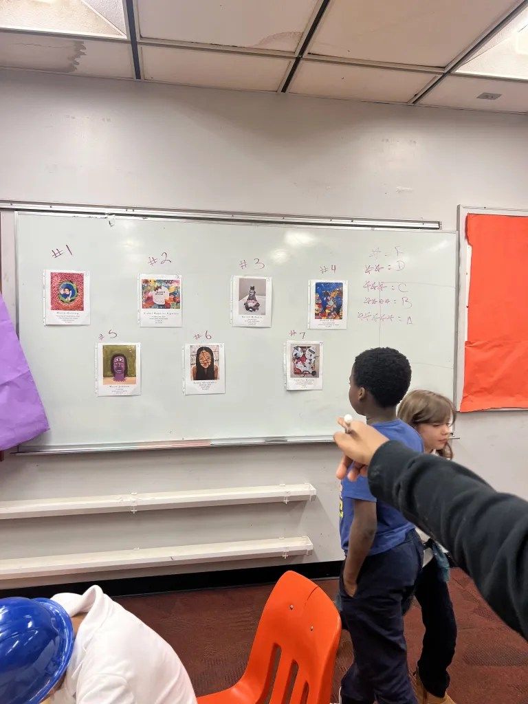 Classroom whiteboard with small portrait cards; two students stand nearby and an orange chair is in front.