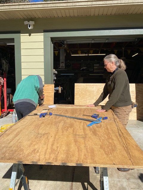 Two people bending over a large plywood board, clamping it outside a garage.