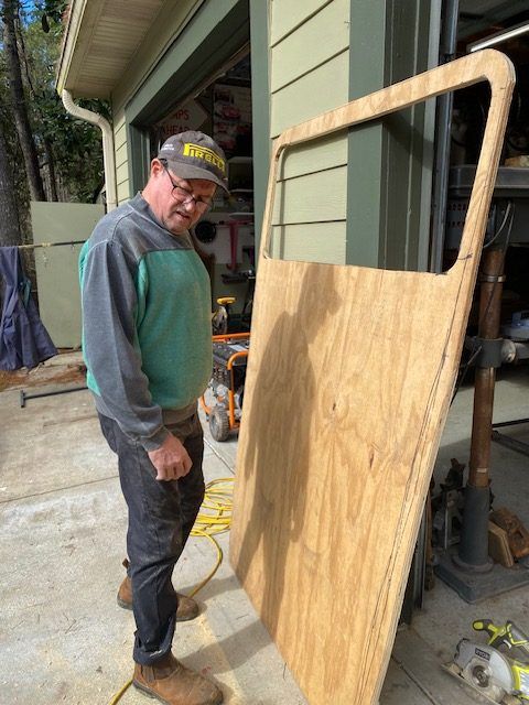 Man in cap standing beside a large plywood panel leaning on a porch