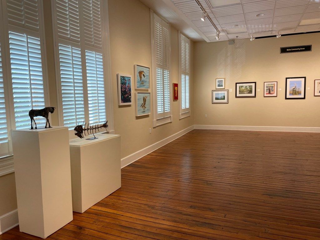 Art gallery room with framed paintings on beige walls and sculptures on white pedestals beside windows