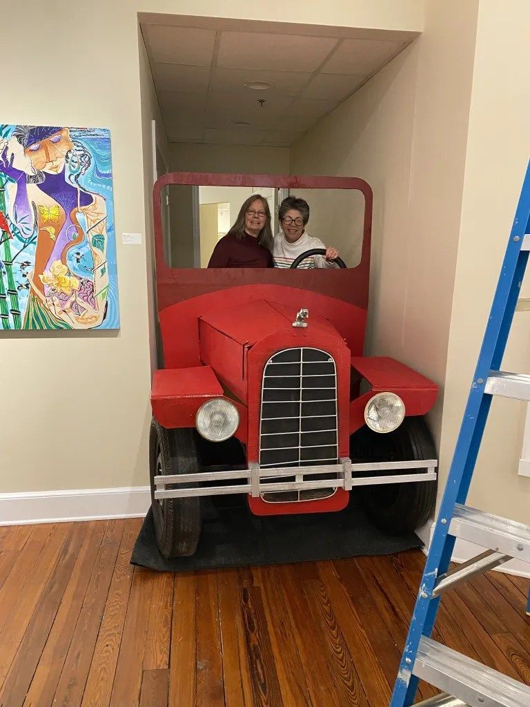 Two people sitting in a red cardboard car display in a hallway, with a painting on the wall nearby.