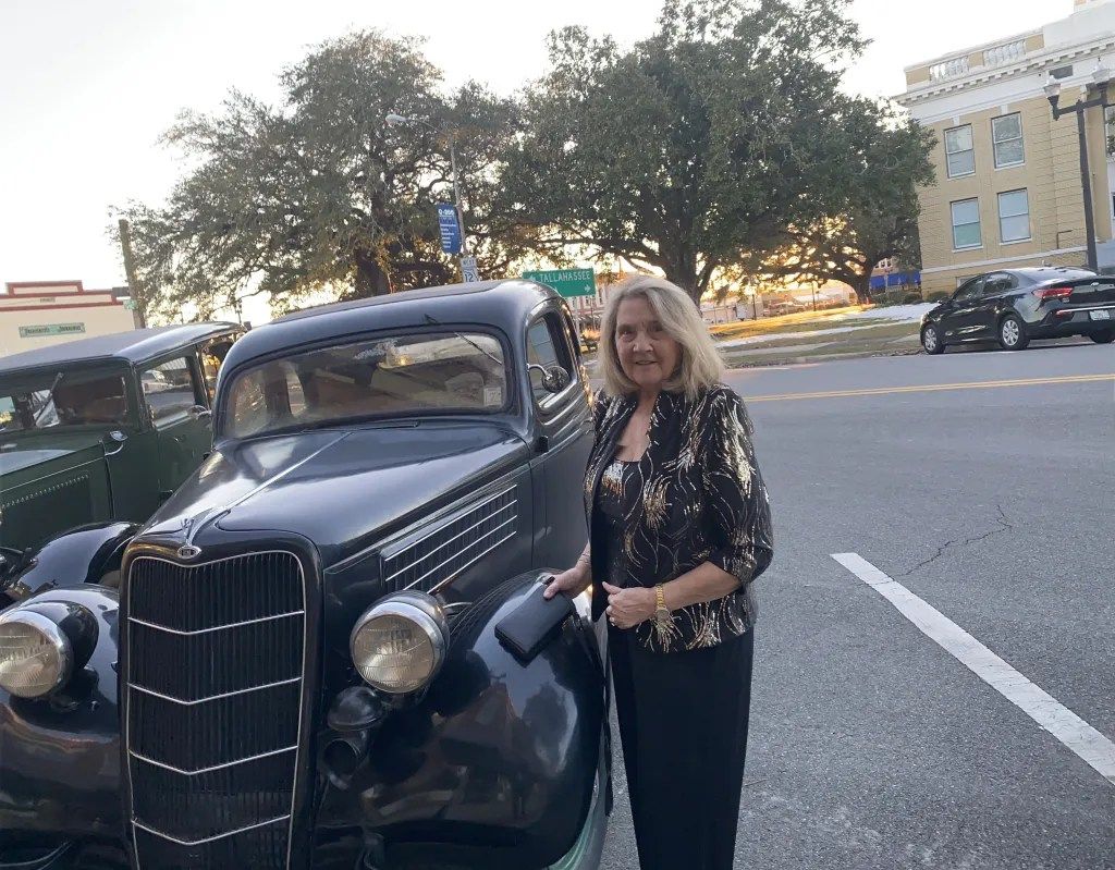 Woman standing beside a vintage black car on a street, with trees and buildings in the background.