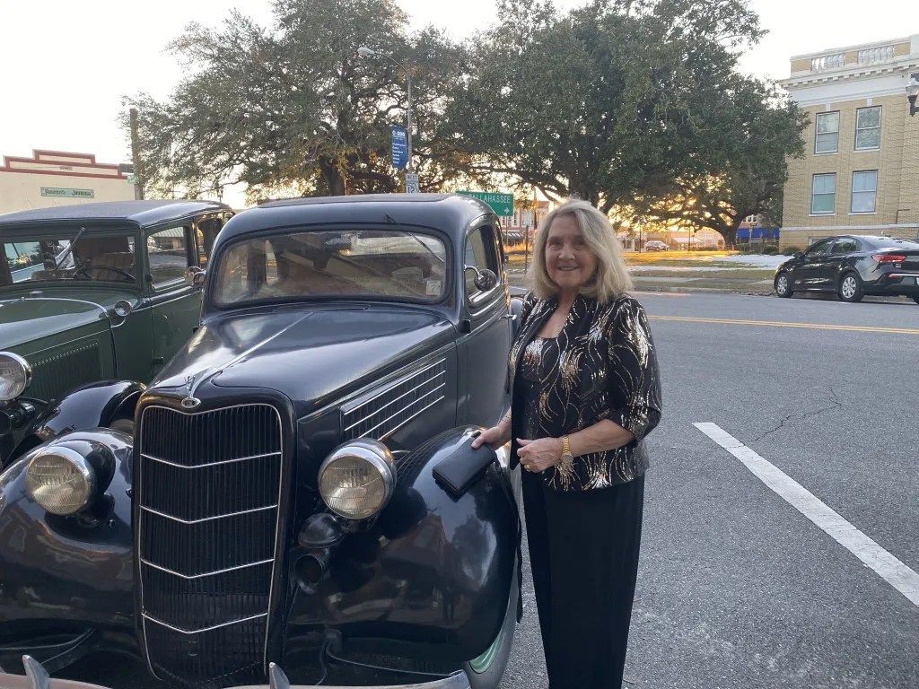 Woman standing beside a vintage black car in a parking lot at dusk