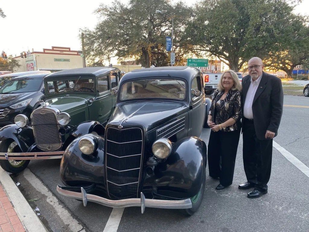 Two people posing beside a black vintage car at a car show on a street at dusk