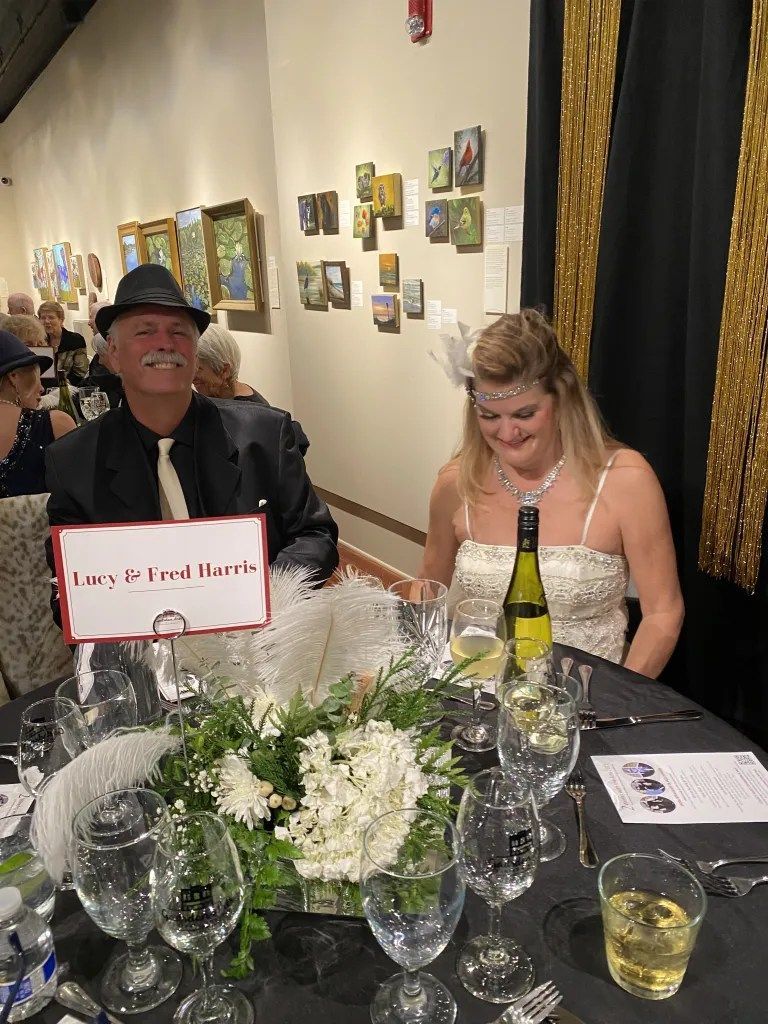 Two people seated at a banquet table with floral centerpiece and drinks, smiling in a warmly lit gallery.