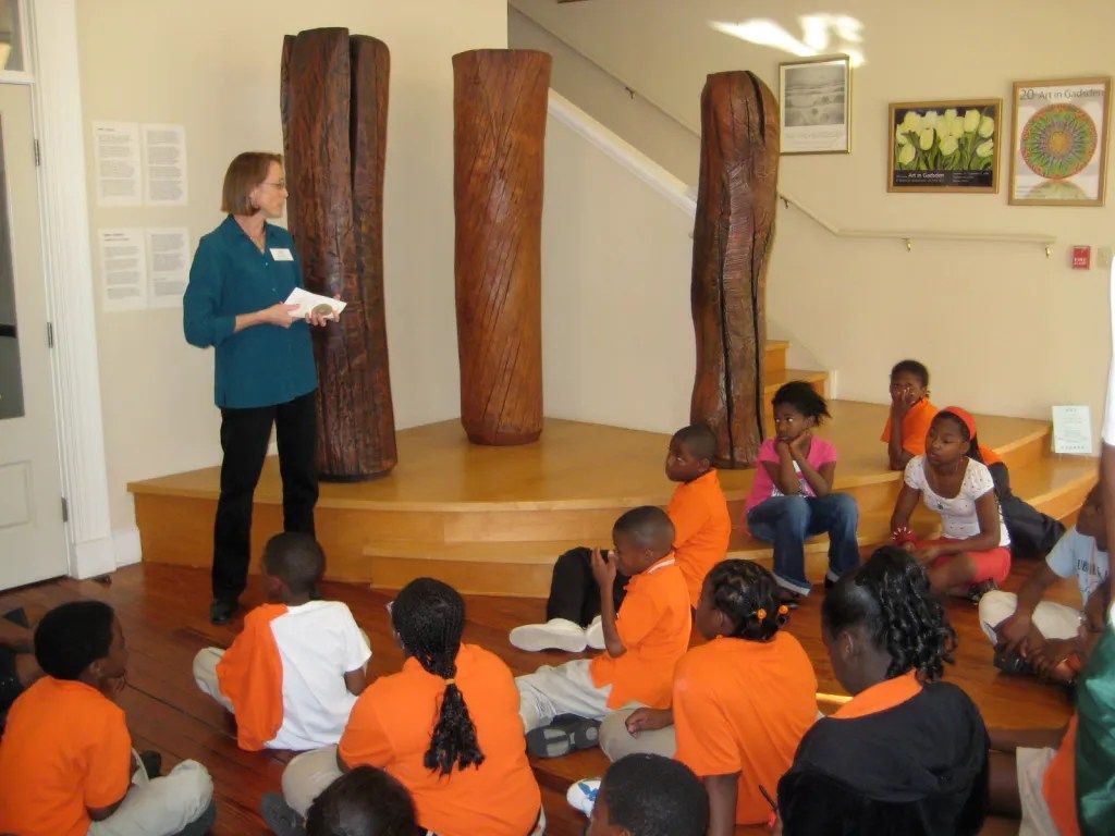 Woman speaking to seated group in a museum exhibit room with carved wooden pillars