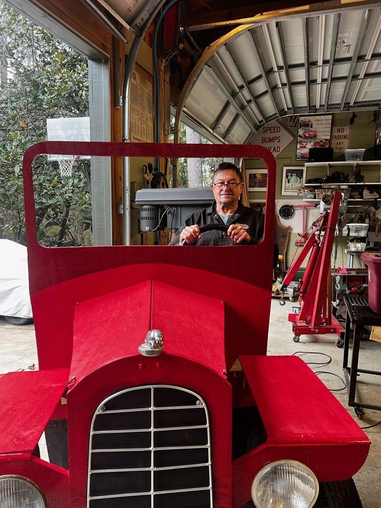 Man standing behind a bright red vintage car frame in a garage, with metal parts and an open door.