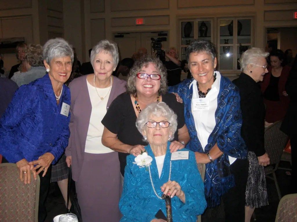 Five women posing indoors at a social event, one seated in front with a cane.