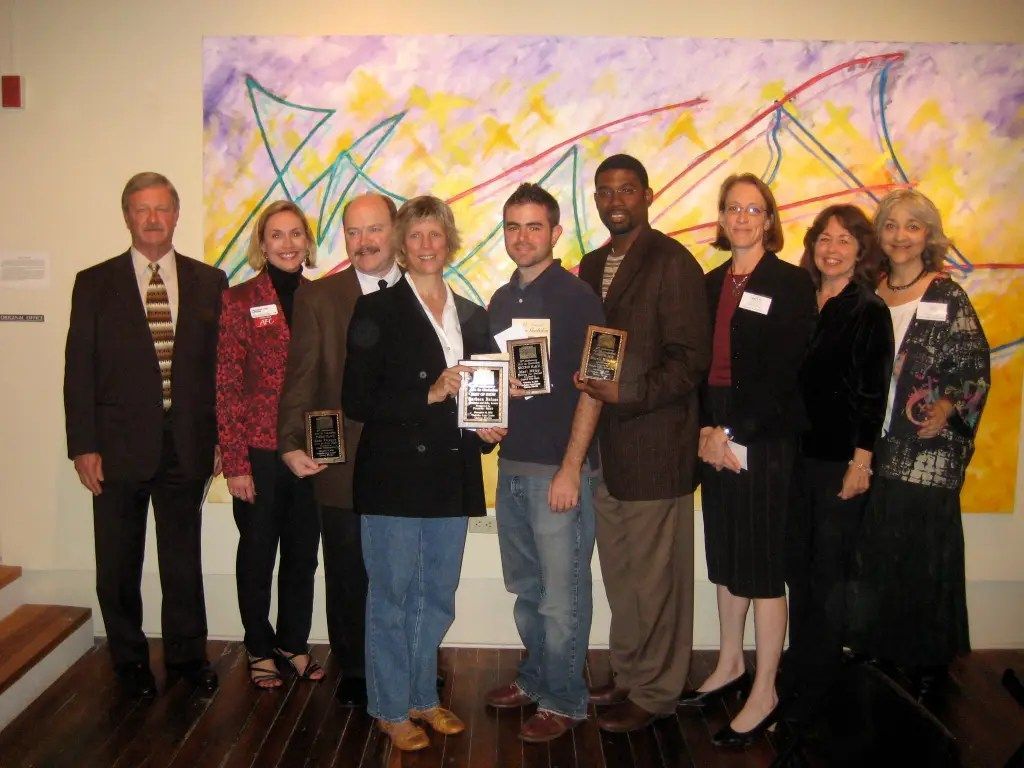 Group of people posing indoors at an awards event, holding framed certificates in front of colorful artwork.