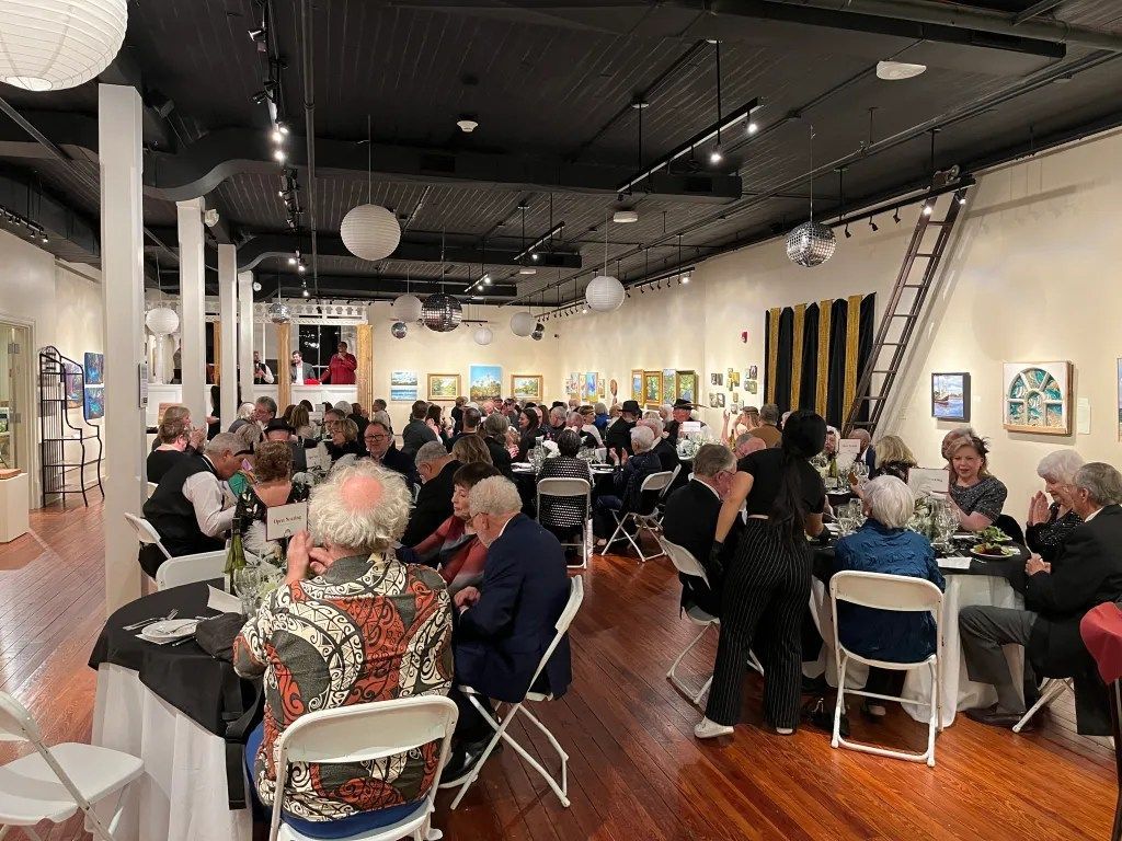 Crowded art gallery with seated guests at tables and framed artwork on bright walls
