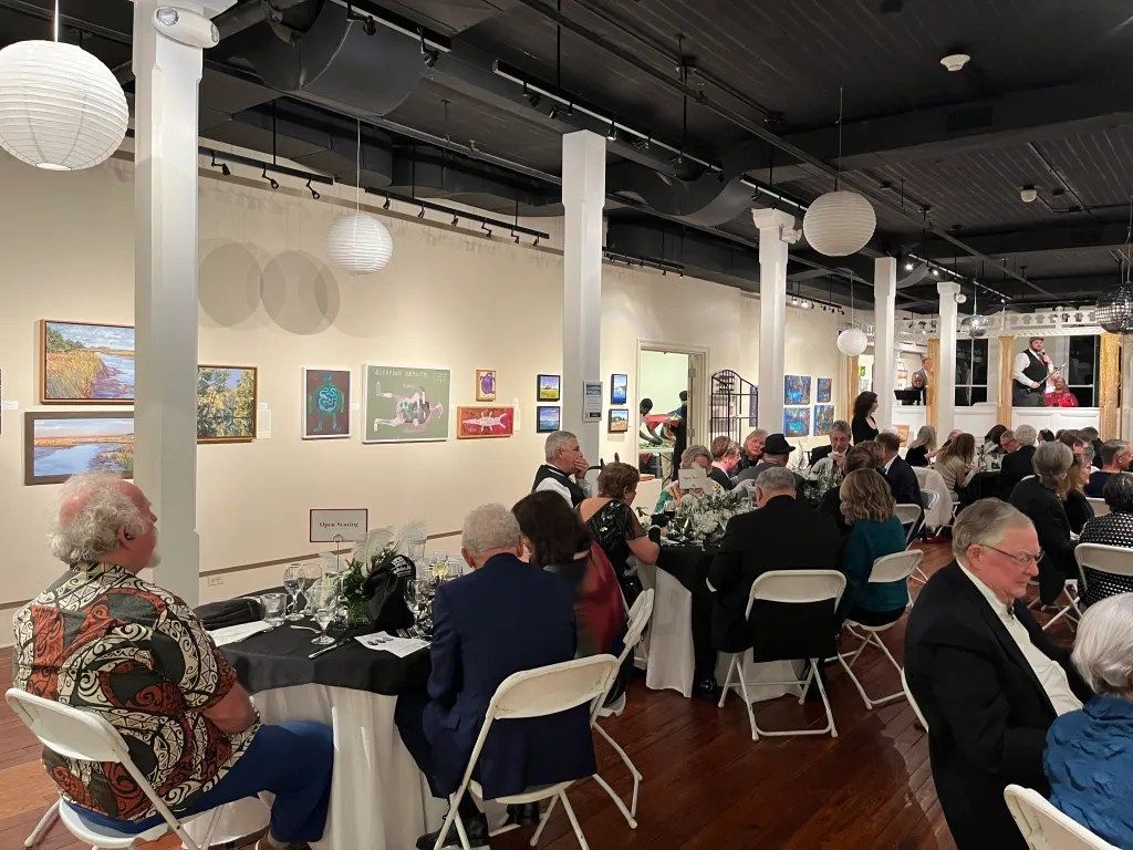 Art gallery with seated crowd at tables, colorful paintings on white walls, and hanging globe lights.
