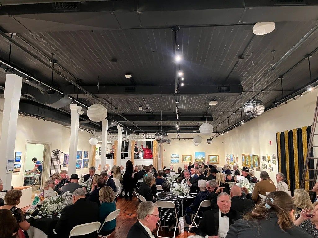 Crowded indoor banquet hall with seated guests at round tables under black ceiling lights and hanging lanterns