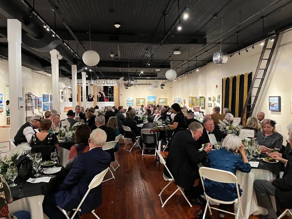 Crowded indoor gallery event with seated guests dining under hanging lights and artwork on the walls