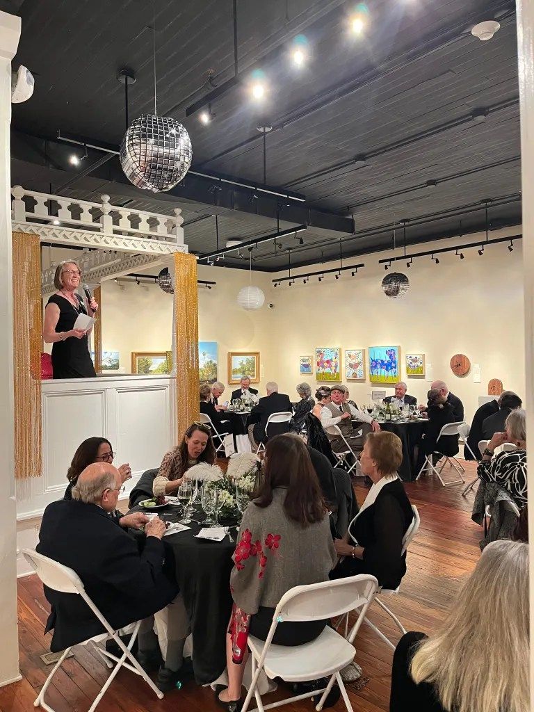 Guests seated at round tables in a gallery-like event space, listening to a speaker onstage under bright lights