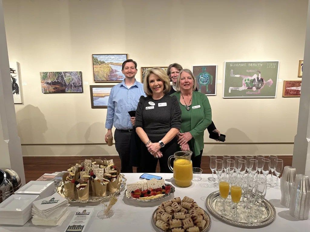 Three people standing behind a dessert table at a gallery event, with framed artwork on the wall.
