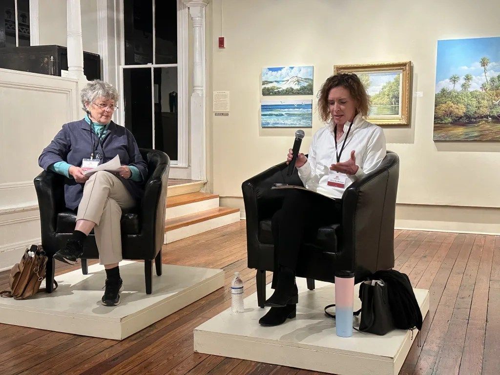 Two people seated in armchairs in a bright gallery or lobby, one taking notes during a conversation.