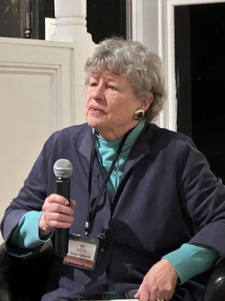 Woman holding a microphone while speaking indoors, wearing a teal shirt and dark sweater.