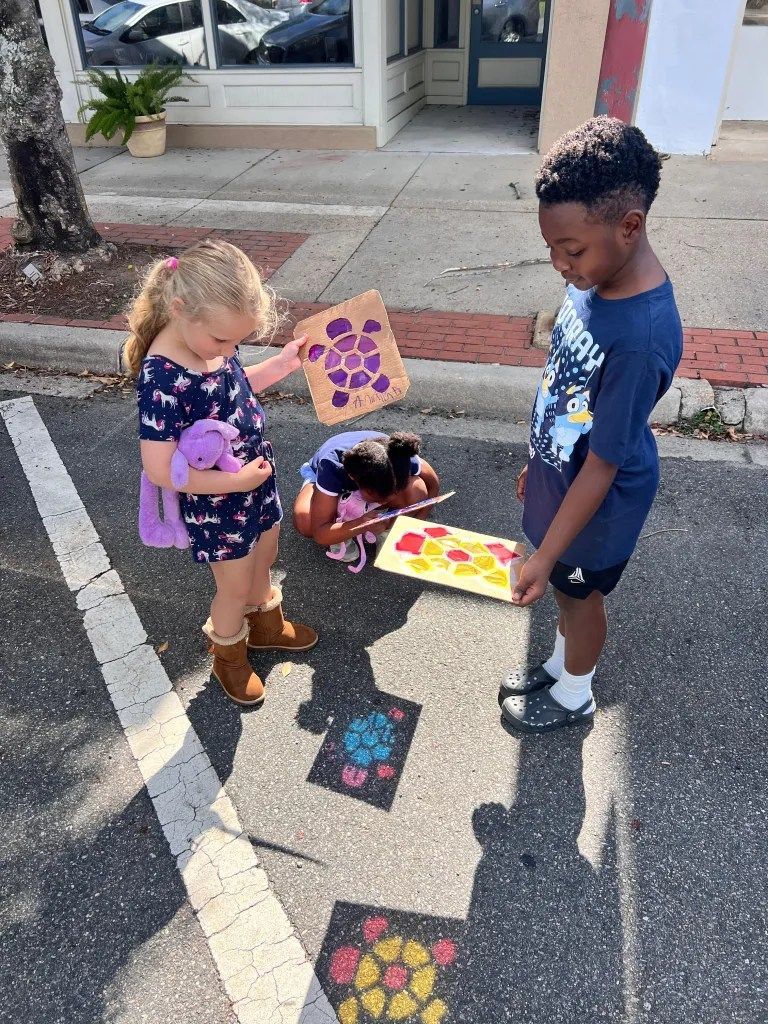 Three children outdoors holding colorful painted paper craft cards on a sunny sidewalk