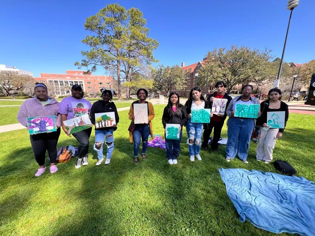 Group of people standing on a grassy field holding certificates, with trees and a blue tarp in the foreground.