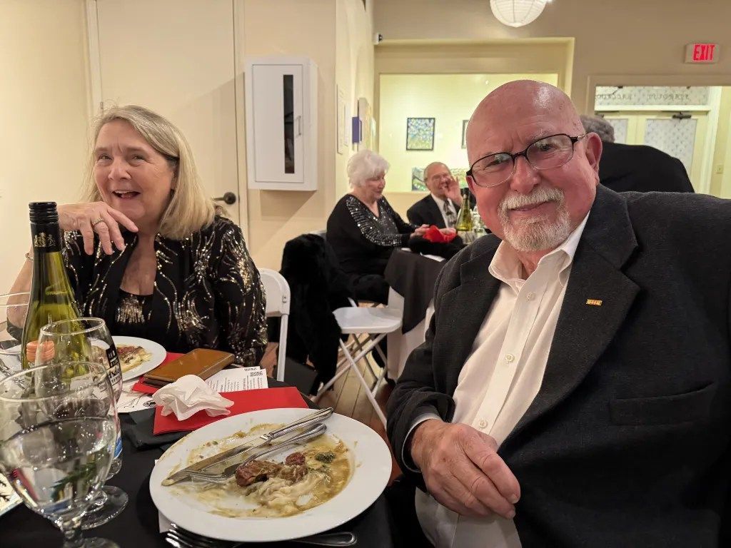 Two people seated at a formal dinner table with wine glasses and plated food, smiling at the camera.