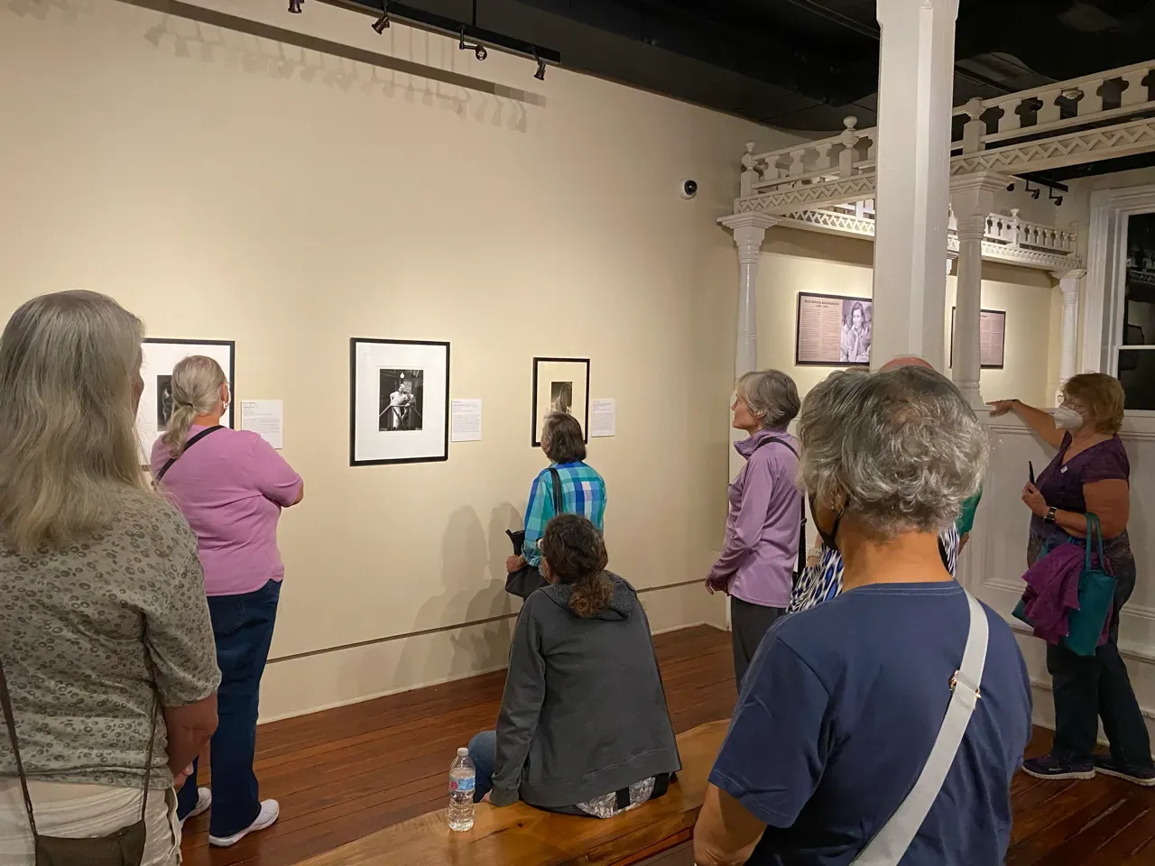People viewing framed artwork in a gallery, with a guide speaking near the wall.