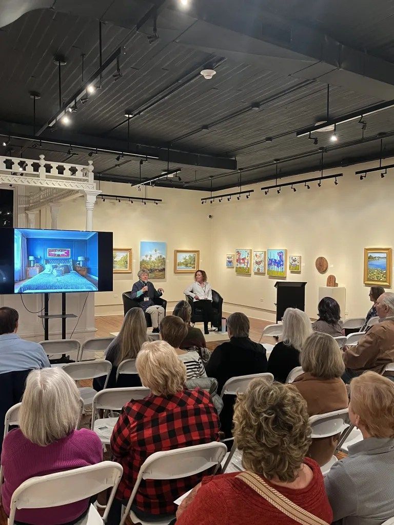 Audience seated in gallery for a talk, watching speakers beside a screen in an art exhibition space.