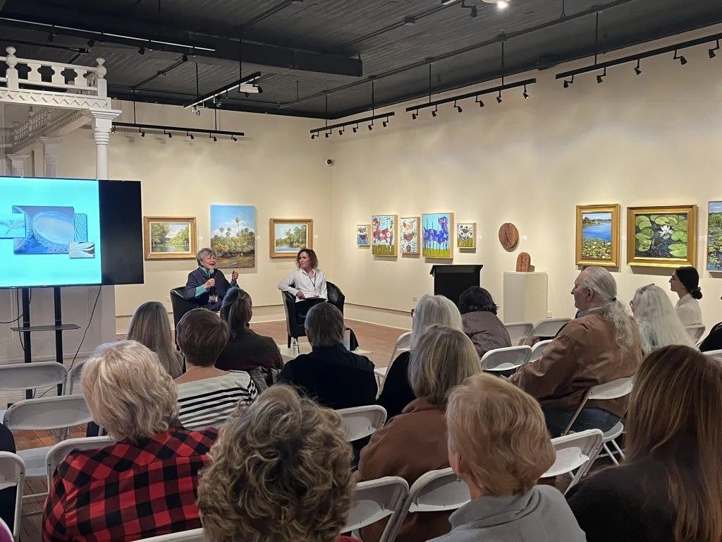 Art gallery talk with seated audience facing two speakers and framed paintings on the walls.