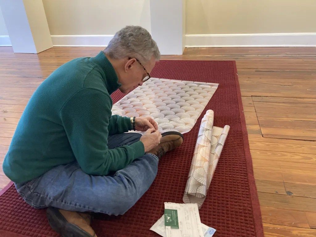 Person kneeling on a red rug, packing a rolled quilt in a living room.