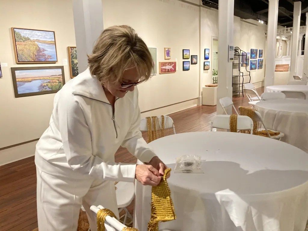 Person in white clothing arranging a gold tablecloth in a gallery with framed art on the walls