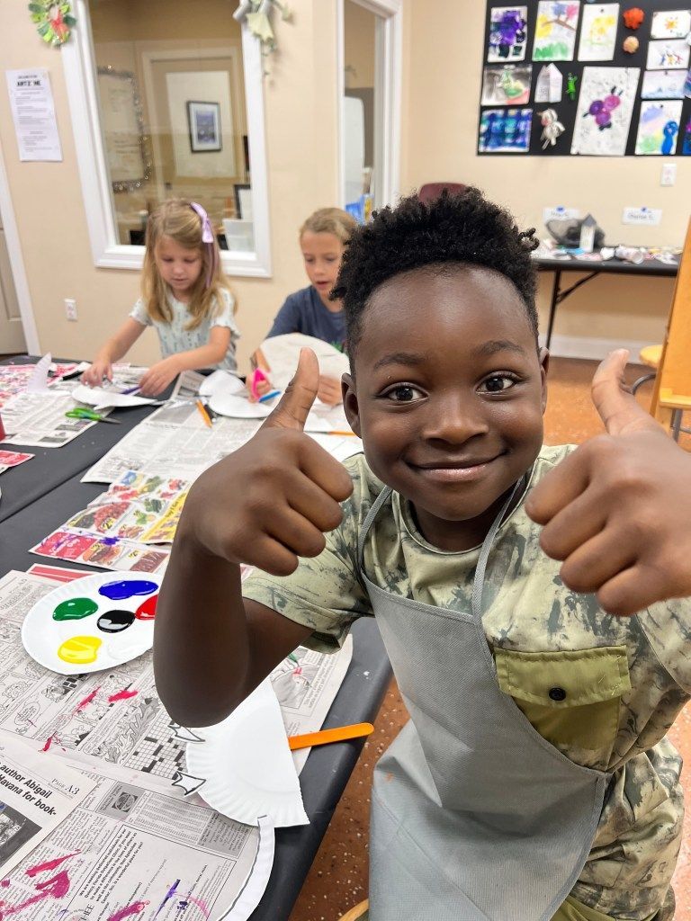 Smiling child giving thumbs-up at an art table with paint, drawings, and other children in the background