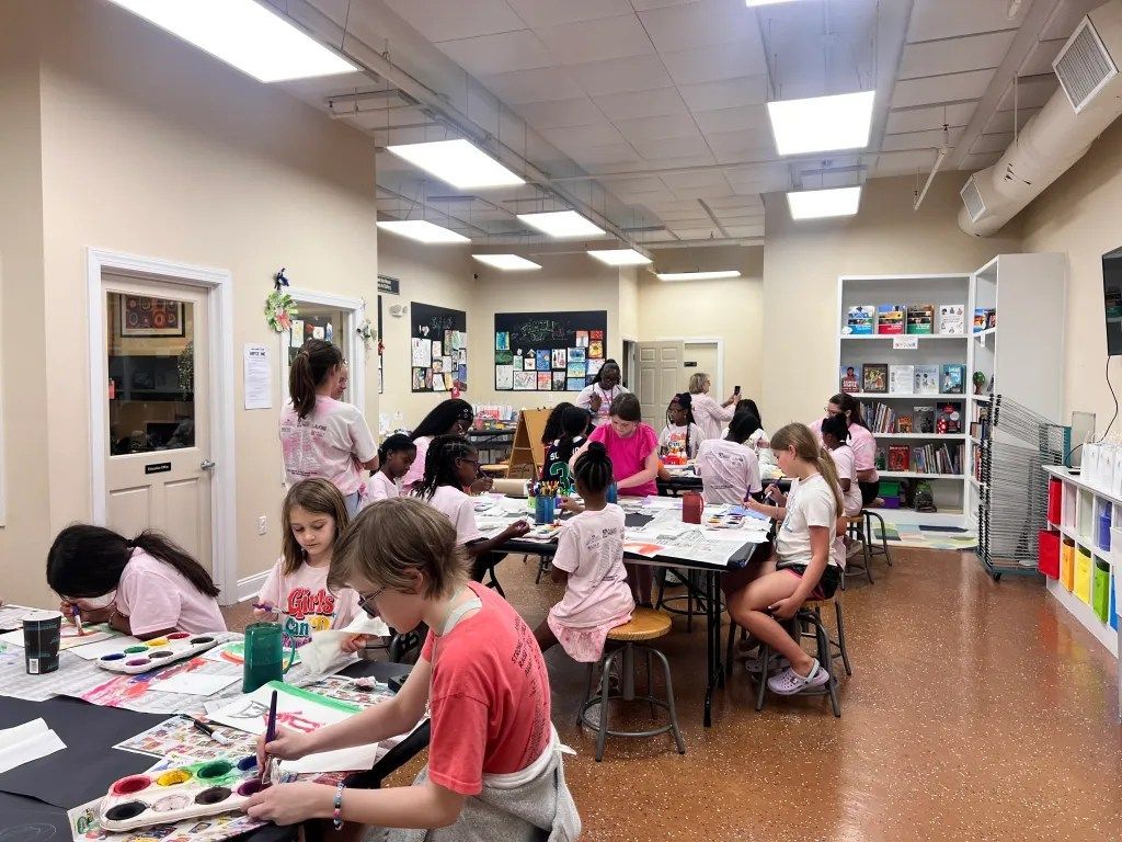 Students painting and drawing at tables in a bright art classroom, with supplies and shelves around them.