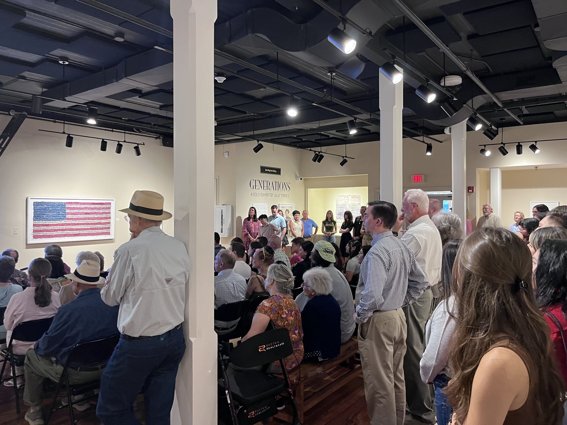 Audience seated in a gallery room, listening to a speaker near an American flag display.