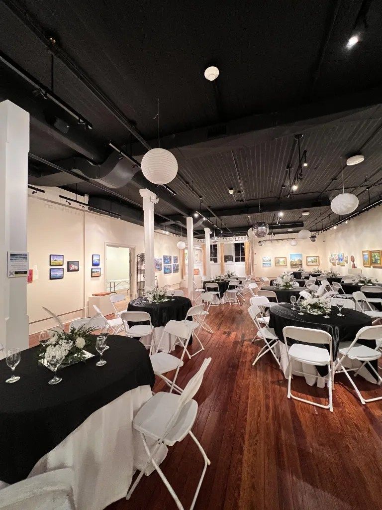 Elegant banquet hall with black ceiling, wood floors, round tables, and white chairs set for an event
