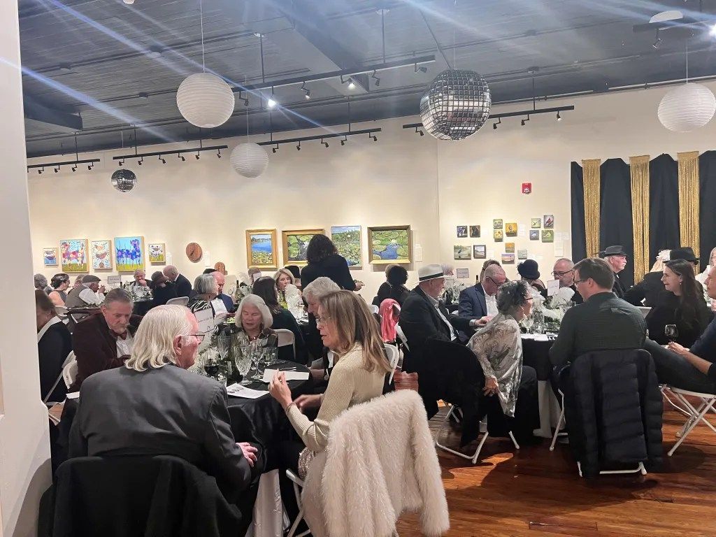 Crowded art gallery reception with seated guests, framed paintings, and hanging globe lights.