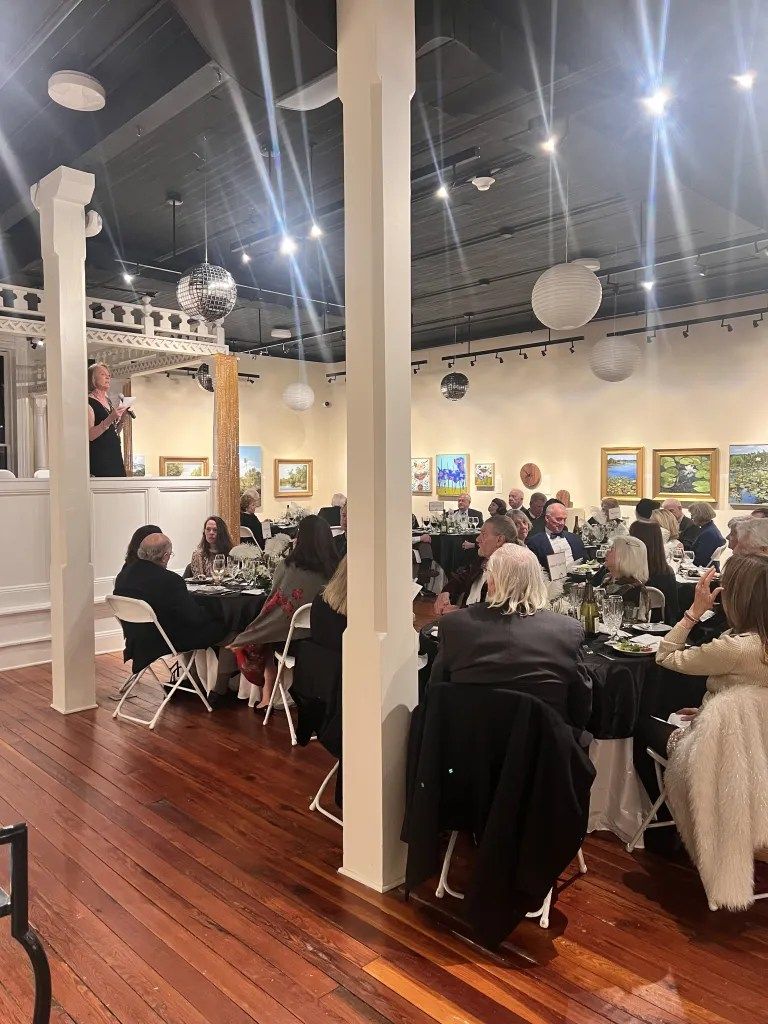 Crowded art gallery event with seated guests, framed paintings, and bright overhead lights