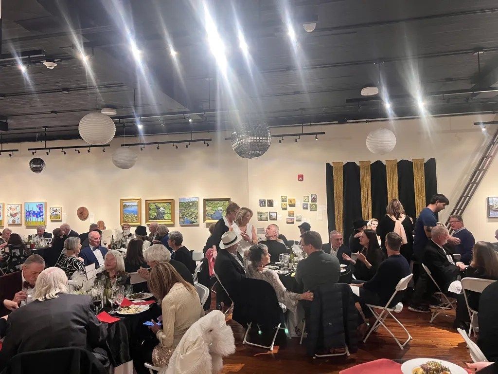 Crowded art gallery event with seated guests, hanging lights, paintings on the walls, and people mingling