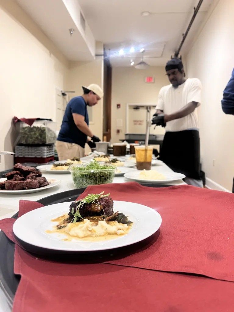 Plated dish on a red tablecloth at a buffet line, with chefs preparing food in the background.