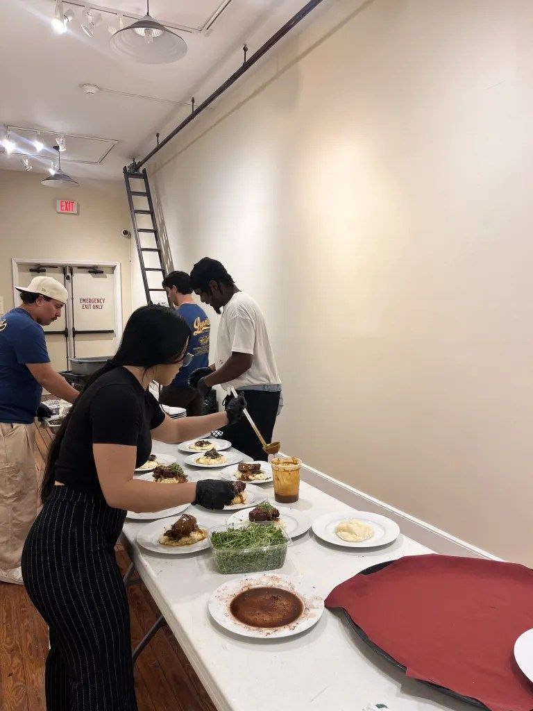 People serving food at a buffet table in a bright hallway, with a ladder against the wall.
