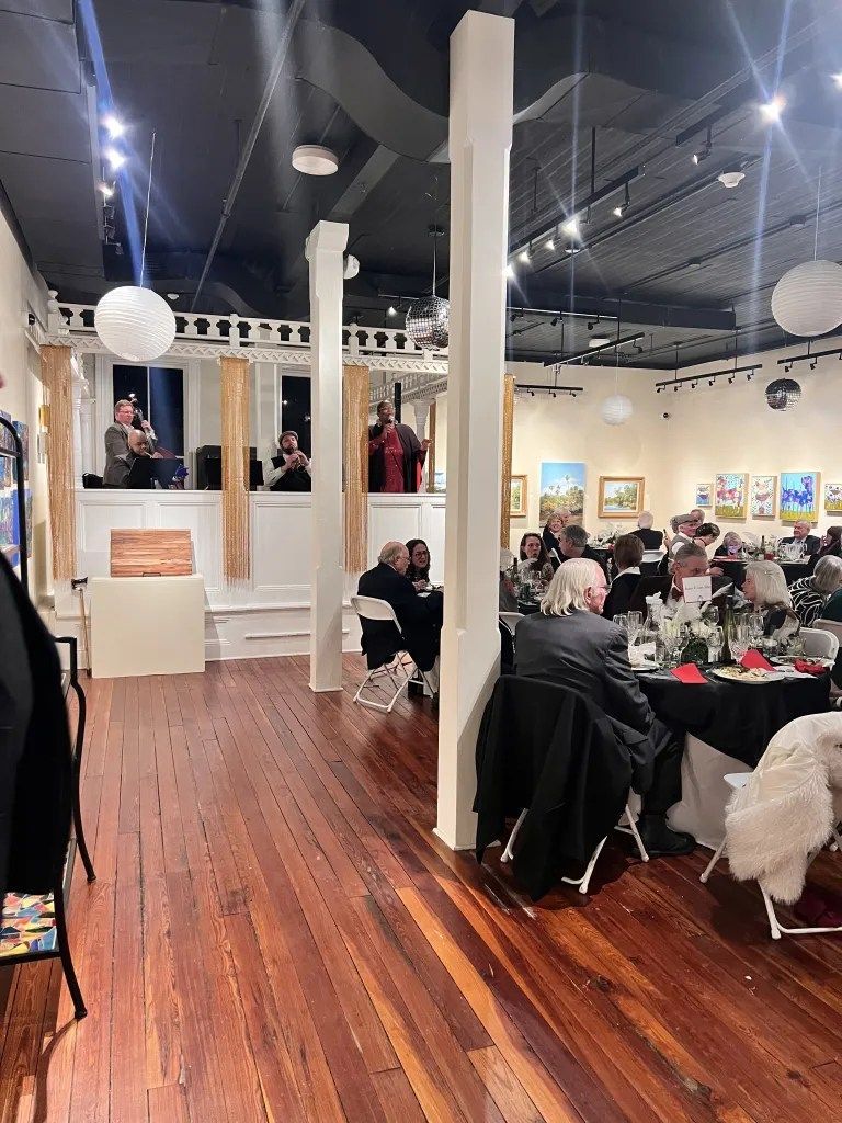 Art gallery event with guests seated at tables in a bright room with wood floors and framed artwork.
