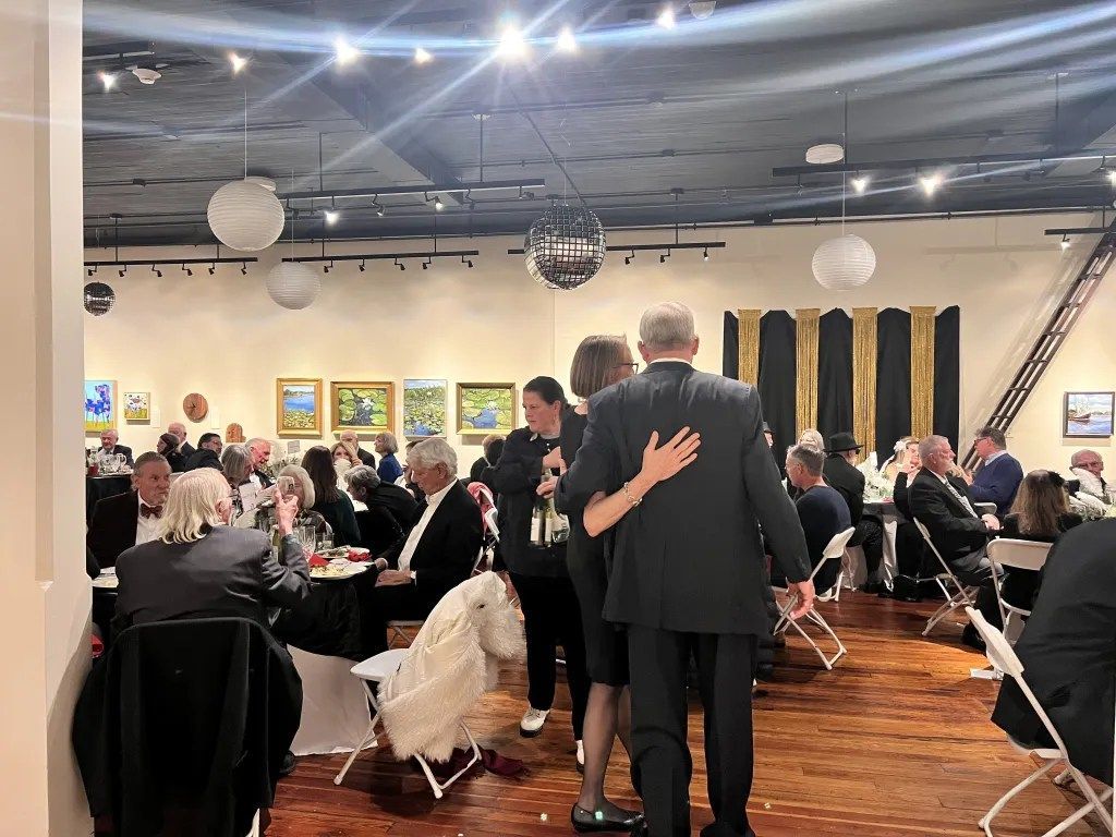 People mingling at a formal indoor reception, with guests seated at tables and two people embracing in the foreground.