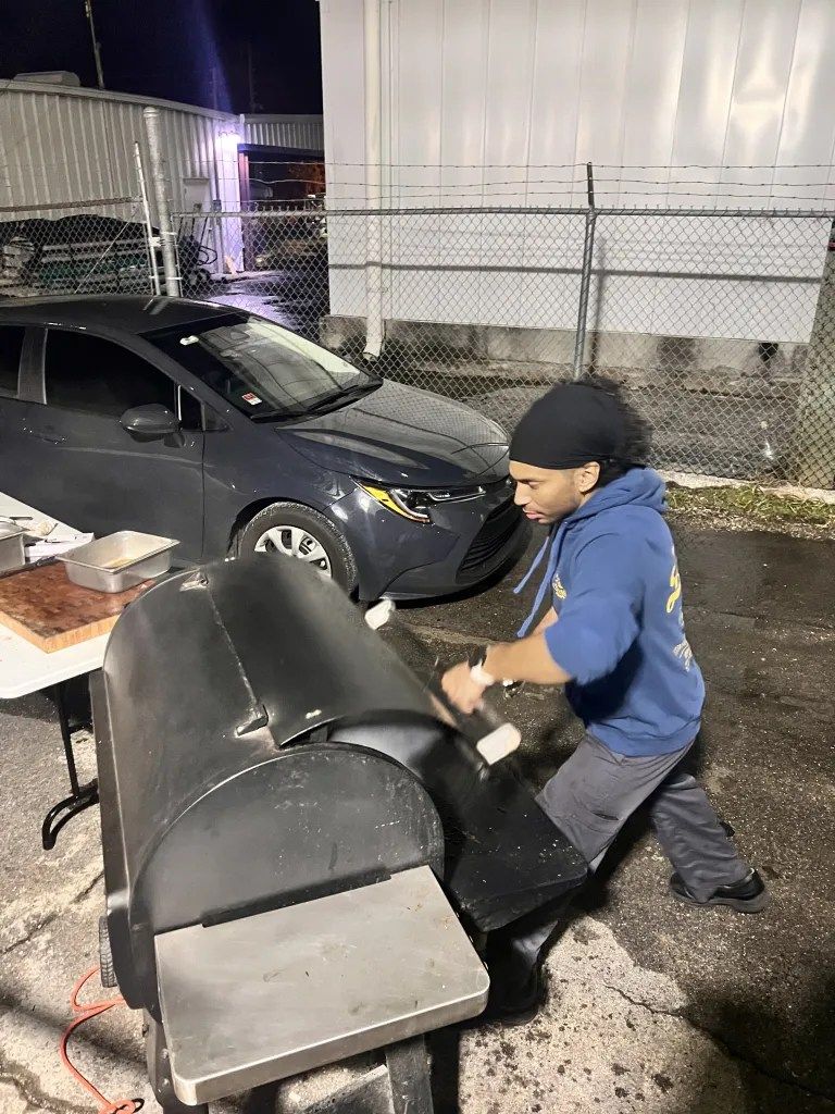 Person pushing a black grill on a wheeled cart beside a parked car at night in a fenced lot