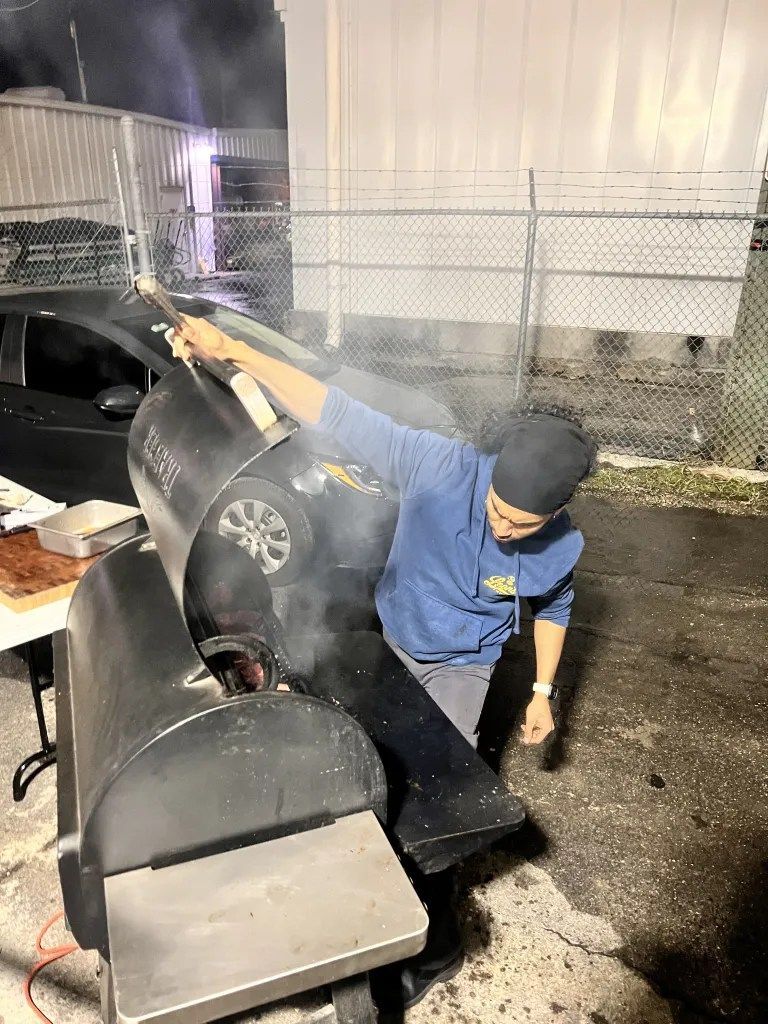 Person in blue jacket beside a smoking grill or appliance in a wet outdoor area at night