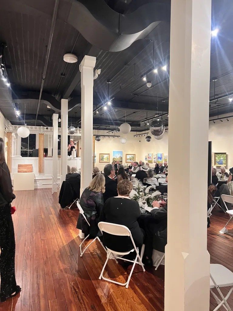 Crowded event hall with seated guests, white columns, dark ceiling, and wood floors
