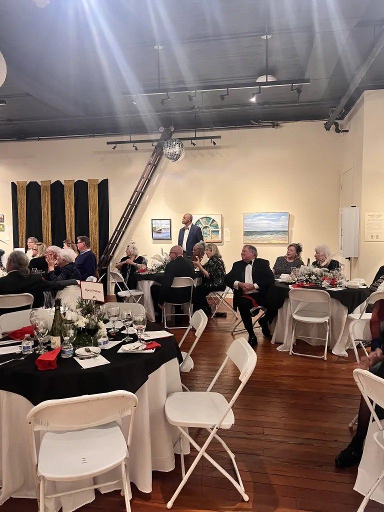 People seated at round tables in a banquet hall with white chairs, black tablecloths, and ceiling beams.