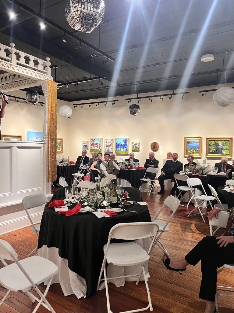 Banquet hall with seated guests at round tables, white chairs, and framed artwork on the walls
