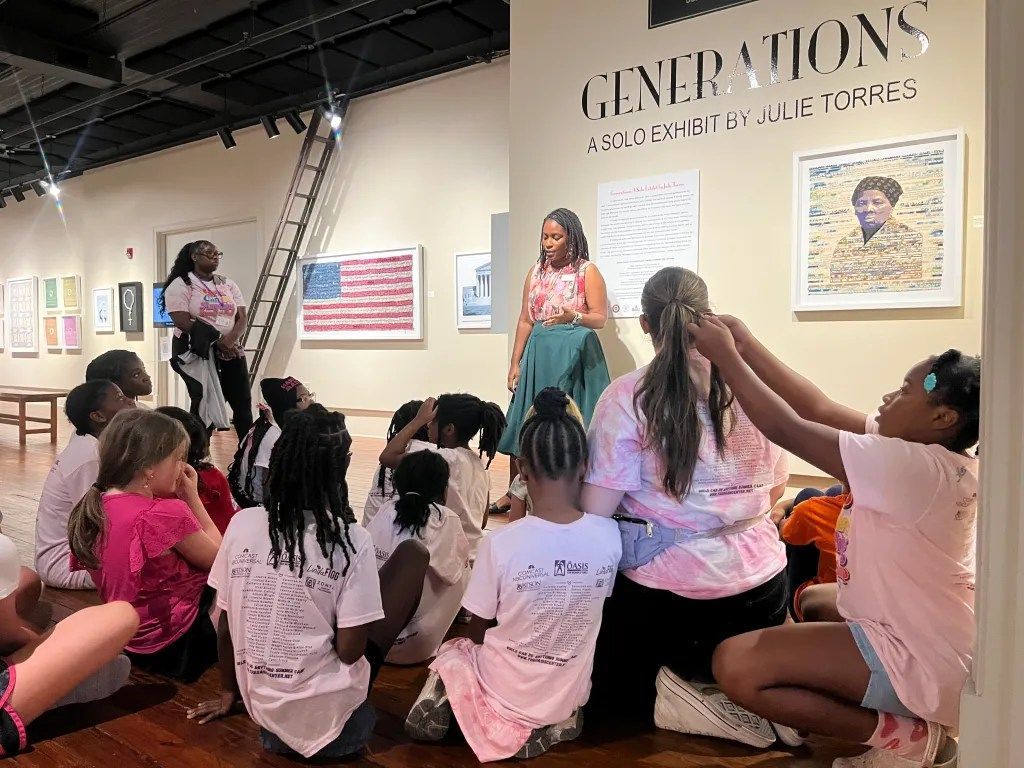 Group seated on floor watching a speaker in a gallery with “Generations” wall art and American flag display