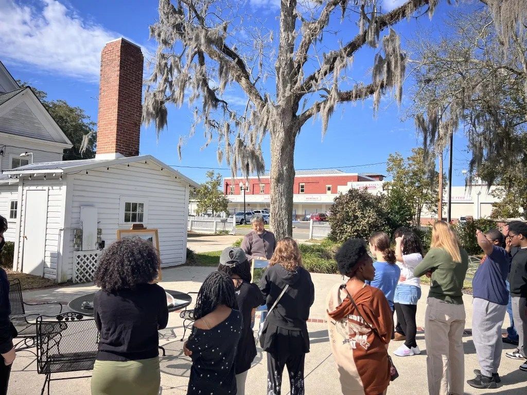 People gathered outdoors near a small white building and brick chimney on a sunny day.